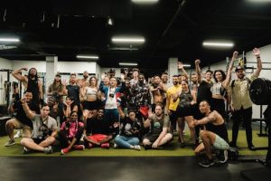 A large group of diverse people smiles and poses energetically in a modern gym setting.
